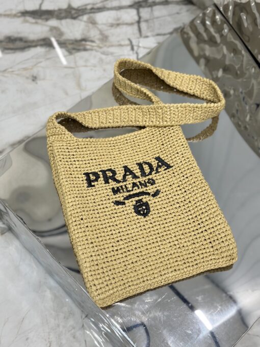 PRADA Crochet Bag- where refined logos meet natural raffia-inspired beauty. Introducing a beautifully crafted tote bag, featuring a gentle, deconstructed design, elegantly crafted from raffia-effect yarn. This lightweight and natural material exudes a delightful summery vibe. The front adorns a sleek enameled metal triangle logo and delicately embroidered lettering logo, engaging in a captivating and harmonious aesthetic conversation.