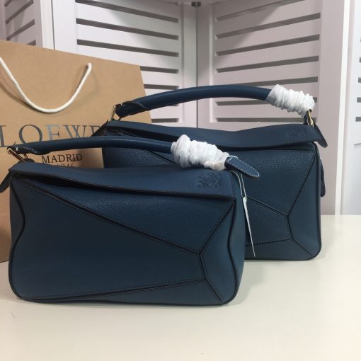 LOEWE 'Puzzle' Shoulder Bag. Authentic quality luxury bag crafted in genuine leather comes with authenticity card, dust bag and gift bag. Unleash the Iconic Cuboid Innovation. The debut bag by Jonathan Anderson has captured the essence of LOEWE and achieved iconic status. This innovative creation showcases a cuboid shape, offering both a remarkable tactile experience and exceptional practicality. The bag's unique volume is achieved through precise cutting, enabling it to be worn in five distinctive styles - from a chic shoulder bag to an elegant clutch - and can even be folded flat for convenient storage. Notable features include the luxurious calfskin craftsmanship, stylish black hand-painted edges, herringbone-patterned cotton-canvas lining, and two internal slip compartments for organized storage. Additionally, a convenient exterior zip back pocket and an embossed anagram add to the bag's functionality and charm. To ensure optimal comfort, the bag also offers a removable adjustable shoulder strap, allowing for a personalized fit.