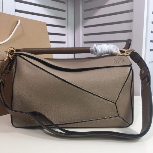 LOEWE 'Puzzle' Shoulder Bag. Authentic quality luxury bag crafted in genuine leather comes with authenticity card, dust bag and gift bag. Unleash the Iconic Cuboid Innovation. The debut bag by Jonathan Anderson has captured the essence of LOEWE and achieved iconic status. This innovative creation showcases a cuboid shape, offering both a remarkable tactile experience and exceptional practicality. The bag's unique volume is achieved through precise cutting, enabling it to be worn in five distinctive styles - from a chic shoulder bag to an elegant clutch - and can even be folded flat for convenient storage. Notable features include the luxurious calfskin craftsmanship, stylish black hand-painted edges, herringbone-patterned cotton-canvas lining, and two internal slip compartments for organized storage. Additionally, a convenient exterior zip back pocket and an embossed anagram add to the bag's functionality and charm. To ensure optimal comfort, the bag also offers a removable adjustable shoulder strap, allowing for a personalized fit.
