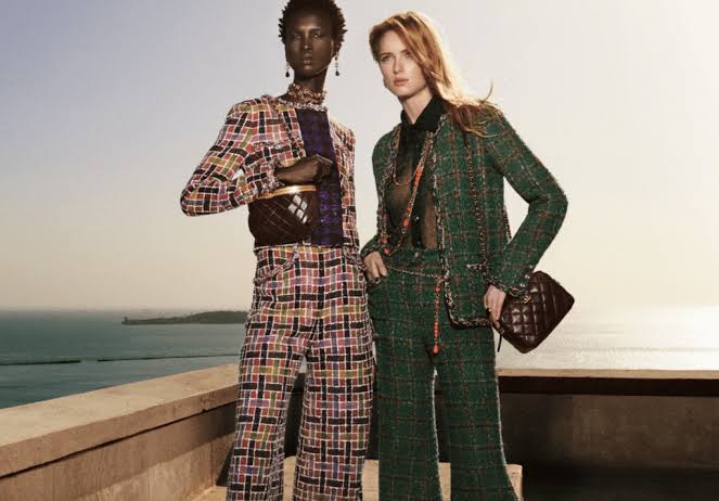 Two models wearing colorful Chanel tweed suits pose confidently on a rooftop with the ocean in the background. Each holds a classic quilted Chanel bag. Two models wearing colorful Chanel tweed suits pose confidently on a rooftop with the ocean in the background. Each holds a classic quilted Chanel bag.
